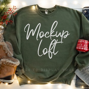 May include: A green sweatshirt with the text "Mockup Loft" in white lettering. The sweatshirt is laid flat on a white surface with a pair of brown boots, a red mug, a gray beanie, and blue jeans.