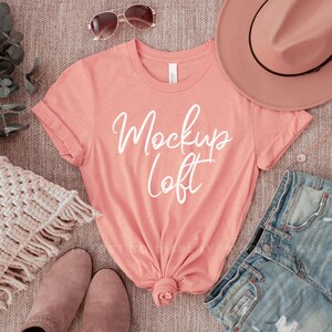May include: A coral pink t-shirt with the text "Mockup Loft" in white. The shirt is laid out on a brown textured surface with a pair of brown boots, a brown hat, a pair of sunglasses, a white crocheted throw pillow, a pair of blue denim shorts, and a silver necklace.