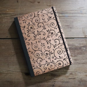 Pode incluir: Um diário de madeira com fecho de banda elástica preta. A capa apresenta um fundo castanho claro com um padrão floral escuro em espiral. A lombada é preta.