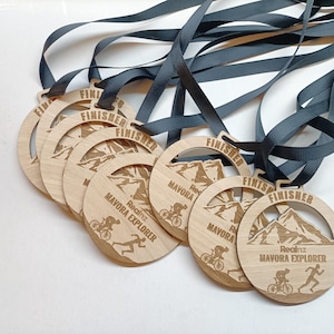 May include: A collection of wooden finisher medals with dark grey ribbons. Each medal displays the word "FINISHER", the text "Realnz Mavora Explorer", and mountain and athlete graphics. The medals are circular with a cut-out.