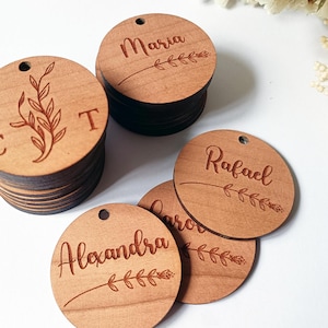 Könnte beinhalten: Ein Stapel runder Holzanhänger mit Namen und einem kleinen floralen Design, das auf jeden Anhänger graviert ist. Die Namen sind Maria, Rafael, Alexandra und CT.