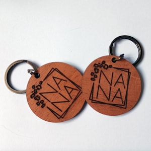May include: Two round wooden keyrings with black metal rings. Each keyring has the word "NANA" engraved in a geometric design.