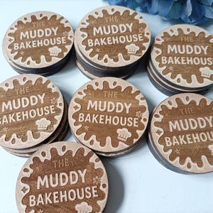 May include: A collection of round wooden coasters. Each coaster features the text "THE MUDDY BAKEHOUSE" in a decorative font, surrounded by a stylized design. The coasters are brown with a lighter brown border and text.