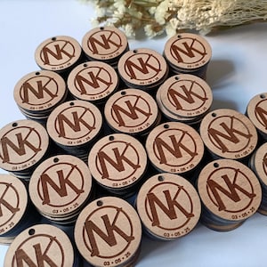 Può includere: Una collezione di targhette rotonde in legno con un monogramma "NK" inciso al laser all'interno di un cerchio. Ogni targhetta include la data "03-05-25". Le targhette sono impilate in piccoli mucchi, mostrando una finitura naturale.
