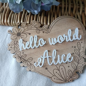 Può includere: Un cartello di legno a forma di cuore con un design floreale. Il cartello dice "hello world" e "Alice" in lettere bianche in rilievo.