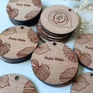 Può includere: Una collezione di ornamenti rotondi in legno con un design a foglie tropicali inciso a laser e la frase "Pura Vida". Alcuni degli ornamenti hanno anche un cerchio con il testo "Circle of Excellence 2021".
