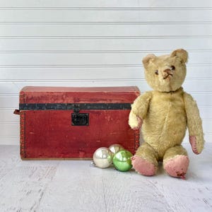 May include: A vintage-style beige teddy bear with pink fabric patches on the paws and feet, positioned next to a rectangular red storage chest with a black metal latch and two round glass ornaments in silver and green colors.