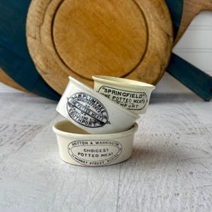 May include: Three antique, off-white ceramic pots stacked on top of each other. Each pot has black text, including "SPRINGFIELD HOME POTTED MEATS", "C.W. PLUMTREE HOME POTTED MEATS", and "BRITTON & WARRINGTON CHOICEST POTTED MEAT". A wooden cutting board is in the background.