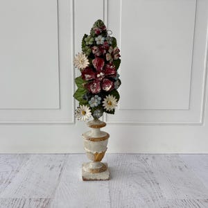 May include: A tall, decorative floral arrangement featuring a variety of artificial flowers in shades of red, white, and blue, atop a cream-colored, ornate pedestal. The pedestal has gold accents and a square base, creating a vintage aesthetic.