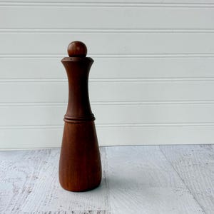 May include: A tall, dark brown wooden pepper mill with a smooth, cylindrical body that tapers toward the center and features a rounded knob at the top. The mill has a wide, stable base and a distinct horizontal ring detail separating the upper and lower sections.