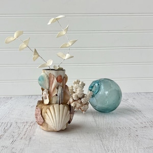 May include: A decorative seashell vase with a variety of seashells and a white, fan-shaped shell. The vase holds a sprig of white, leaf-shaped stems. A light blue glass float and a piece of white coral are next to the vase.