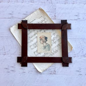 May include: A dark brown wooden picture frame with carved leaf details at each corner. The frame holds a small, rectangular print featuring a profile illustration of a person with reddish hair, set against a sheet of vintage musical notation paper.