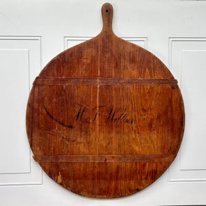 May include: A large, round, brown wooden bread board featuring a handle at the top and two horizontal wooden support slats across the front. The surface displays a visible wood grain and the handwritten black ink inscription H.T. Walker.