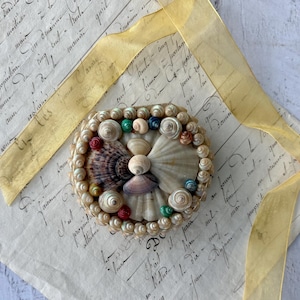 May include: A circular seashell art piece featuring a variety of shells in shades of white, brown, and beige. The shells are arranged in a decorative pattern, with colorful accents of red, green, and blue. A gold ribbon and handwritten script background complete the design.