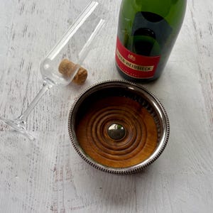 May include: A champagne flute with a cork, a wooden coaster with a silver rim, and a green champagne bottle with a red label. The coaster has a wooden center with concentric circles and a silver metal rim.