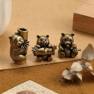 May include: Three bronze panda figurines. One panda is hugging a bamboo stalk, another is holding a bamboo log, and the third is holding a bamboo stick. The pandas have detailed features and a vintage finish. The figurines are displayed on a brown surface.