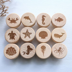 Peut inclure: Ensemble de 12 tuiles rondes en bois avec divers animaux marins et objets gravés dessus. Les tuiles sont de couleur brun clair et les gravures sont de couleur brun foncé. Les animaux marins comprennent un crabe, un dauphin, un hippocampe, un poisson, une tortue, une étoile de mer, une coquille, une sirène, une méduse et une pieuvre.