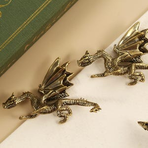 Peut inclure: Tirants de meubles en forme de dragon en laiton antique. Ces tirants décoratifs présentent des figurines de dragon détaillées avec des ailes déployées. La quincaillerie est de couleur or chaud, parfaite pour ajouter une touche de fantaisie.