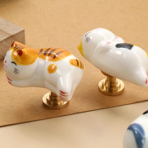 Könnte beinhalten: Zwei keramische Schrankknöpfe in Katzenform. Einer ist weiß mit schwarzen und gelben Markierungen, der andere ist orange und weiß mit Tigerstreifen. Beide haben messingfarbene Sockel und liegen. Die Knöpfe befinden sich auf einer hellbraunen Oberfläche.