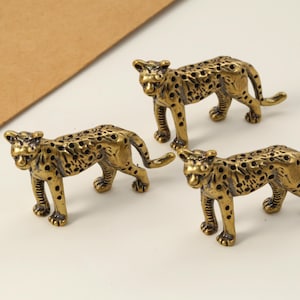 Puede incluir: Tres pequeñas figuras de leopardo doradas con manchas negras. Las figuras están en posición de caminar, con las colas curvadas. El fondo es una superficie clara con un papel marrón.