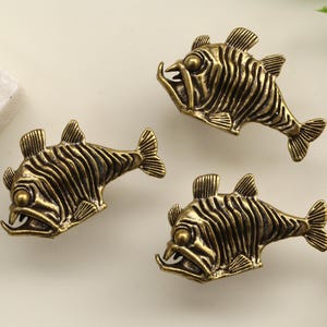 May include: Antique brass fish-shaped cabinet knobs. The knobs have a detailed, textured design with fins, scales, and open mouths. Suitable for furniture or other home decor projects. The knobs are in a warm, antique brass colour.