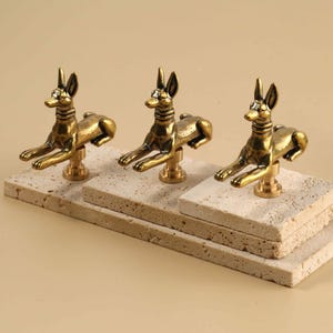 May include: A set of four brass-coloured metal finials, each shaped like a reclining jackal or dog with pointed ears and a collar. These decorative finials are mounted on small, square, light-coloured, porous stone bases arranged in a tiered formation.