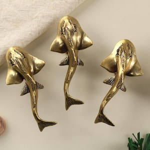 May include: Three brass-colored, stylized rayfish sculptures. Each sculpture has a flat, wing-like body and a long, tapering tail. The rayfish are arranged on a neutral surface, with a seashell and a piece of greenery nearby. The sculptures are likely decorative items.