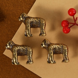 Könnte beinhalten: Drei bronzefarbene Gorilla-Figuren sind auf einer braunen und beigen Oberfläche angeordnet. Die Gorillas stehen. Ein Zweig mit roten Beeren befindet sich in der oberen rechten Ecke. Der Hintergrund ist eine Mischung aus Braun und Beige.