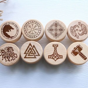May include: Eight wooden round objects with engraved Viking symbols. The symbols include a wolf, a compass, a cross, two birds, a horse, a triangle, a hammer, and a hammer with a triangle. The objects are light brown and arranged in two rows of four.