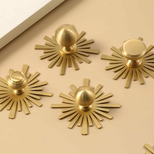 May include: Four gold-colored metal cabinet knobs, each featuring a central circular knob mounted on a flat, sunburst-shaped backplate with radiating rays. The hardware has a smooth, matte finish and is arranged in a group on a flat surface.