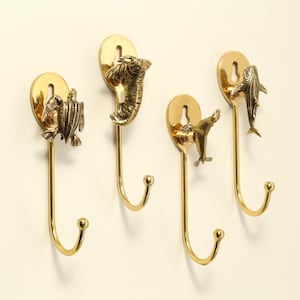 Könnte beinhalten: Vier Messing-Wandhaken mit Tiermotiven: eine Schildkröte, ein Elefant, eine Robbe und ein Fisch. Jeder Haken hat eine dekorative Tierfigur, die an einer ovalen Messing-Rückplatte und einem gebogenen Haken befestigt ist.