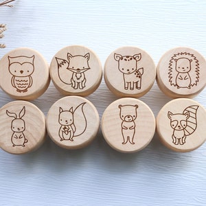 May include: Eight wooden round blocks with engraved woodland animal designs: an owl, fox, deer, hedgehog, bunny, squirrel, bear, and raccoon. The blocks are light brown and arranged in two rows of four.