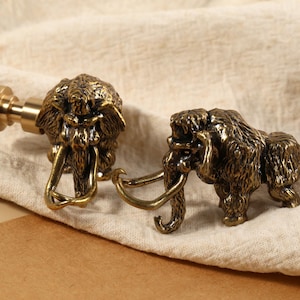 Peut inclure: Deux figurines de mammouths de couleur bronze. L'une est fixée à une tige, l'autre est debout. Les mammouths ont des fourrures et des défenses détaillées. Le fond est un tissu beige.