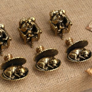 May include: Six brass decorative drawer pulls. Three pulls depict a figure seated in a chair, and three pulls depict a sleeping dog in a shell. The pulls have a vintage, antique aesthetic.