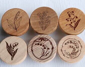 May include: Six round wooden coasters with laser-etched lily of the valley designs. Each coaster features a different arrangement of the flowers.