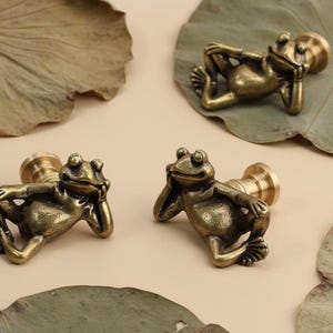 May include: Four antique brass frog-shaped cabinet knobs. Each frog is in a relaxed pose, resting its head on its hand. The knobs have a cylindrical base. The background includes dried leaves and a neutral-toned surface.