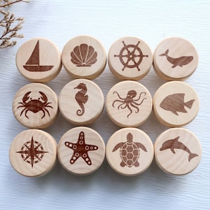 May include: Twelve wooden cabinet knobs with nautical designs. Each knob features a different engraved image, including a sailboat, seashell, ship's wheel, whale, crab, seahorse, octopus, fish, compass, starfish, turtle, and dolphin.