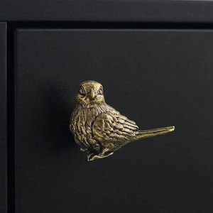 May include: A brass-coloured metal cabinet knob shaped like a small bird with detailed feather textures. The bird is depicted in a side profile, facing forward, and is mounted onto a flat, matte black surface, serving as a functional handle.