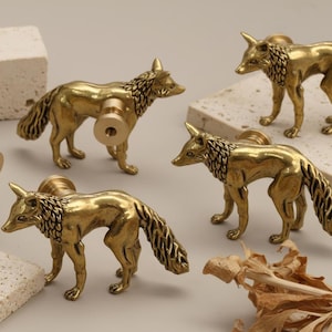 May include: A selection of brass fox-shaped cabinet knobs. The knobs feature detailed fur and tails, and are presented on neutral blocks. The foxes are in different poses, some with the knob hardware attached.