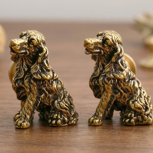 May include: Two identical brass-toned cabinet knobs shaped like sitting spaniel dogs. Each knob features detailed fur texture, floppy ears, and a forward-facing posture. The metallic finish has darkened highlights in the crevices to emphasize the sculptural form.