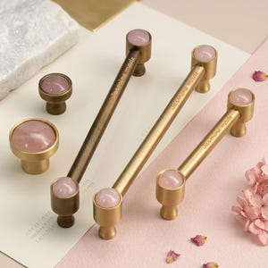 May include: Three brass cabinet pulls and two round knobs featuring inlaid pink rose quartz stone accents. Each handle is engraved with the text Jasmine made. The hardware has a brushed metallic finish and cylindrical mounting posts.