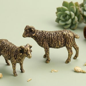 Peut inclure: Deux figurines de bélier en bronze avec des textures détaillées se tiennent sur une surface vert clair. Les figurines de bélier sont positionnées près d'un petit livre vieilli et d'une plante succulente. Des boutons de fleurs séchées sont éparpillés.