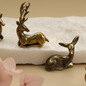 May include: Several small, antique brass deer figurines in various poses. The deer have detailed antlers and are resting on a white surface. Soft pink flowers are in the foreground, adding a touch of color to the scene.