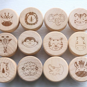 May include: Twelve wooden animal toys, each with a different engraved animal face. The collection includes a giraffe, lion, elephant, tiger, monkey, hippo, rhino, sheep, crocodile, and zebra. The toys are made of light-coloured wood and are round in shape.