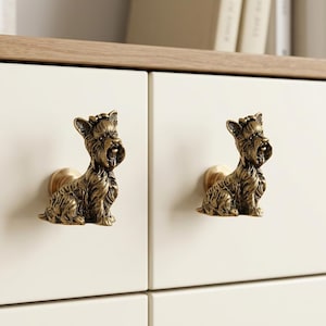 May include: Two antique brass-toned cabinet knobs shaped like sitting Yorkshire Terrier dogs. Each knob features detailed fur texture and a small bow on the head, mounted on a circular base attached to a smooth, off-white cabinet drawer front.