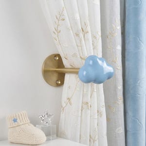 Peut inclure: Une embrasse de rideau murale composée d'une base circulaire et d'une tige horizontale en métal doré, terminée par un bouton bleu clair brillant en forme de nuage. Elle retient un rideau blanc brodé de motifs de feuilles et de nuages dorés.