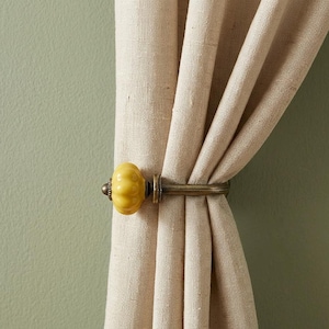 May include: A beige fabric curtain held in place by a metal curtain tieback featuring a round, yellow, pumpkin-shaped ceramic knob with a textured surface and a dark bronze-toned metal base and arm that secures the fabric in a gathered fold.