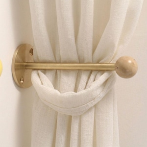 May include: A brass curtain tieback featuring a circular wall-mounted base with two screw holes, a horizontal rod, and a smooth spherical end piece. The metal hardware holds a gathered off-white linen-textured curtain fabric in place.