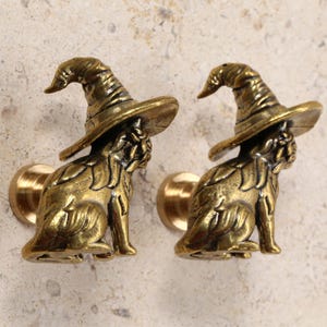 May include: Two antique brass cabinet knobs. Each knob features a sitting dog wearing a witch's hat. The knobs are a warm gold color and are mounted on a round base. The background is a light beige.