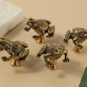 Può includere: Quattro pomelli per mobili a forma di rana in bronzo. Ogni pomello presenta un design dettagliato a forma di rana con una base in ottone. Le rane sono in varie pose, aggiungendo un tocco stravagante a mobili o decorazioni.